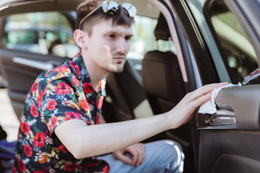 Portrait of one young Caucasian handsome brunette man wiping black leather upholstery of rear passenger door of white car with wet cloth on sunny spring day at city car wash, side view very close-up with selective focus. Concept time car wash, working people.