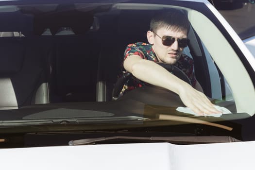 Portrait of one young Caucasian handsome brunette man in sunny, who wipes with a wet cloth the black dashboard of a white car on a sunny spring day at a city car wash, side view medium shot. Concept time car wash, working people.