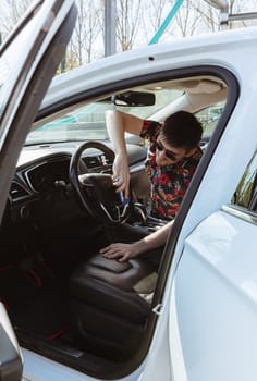 Portrait of a young handsome Caucasian man wiping the steering wheel of a car with a special sponge while sitting in the passenger seat on a sunny spring day at a city car wash, side view, medium shot. Concept: time to wash a car, working people.