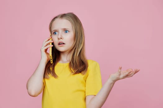 A young girl with long hair dressed in a bright yellow shirt is surprised while talking on the phone, set against a soft pink background, showcasing emotions of confusion and curiosity