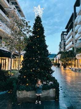 Little smiling girl stands near a Christmas tree decorated with luminous stars on top. High quality photo