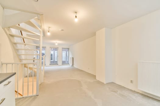 A spacious, minimalist indoor area featuring a staircase and ample natural light. The room has a neutral palette and unfinished flooring, offering a blank canvas.