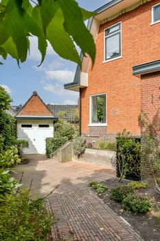 A picturesque scene featuring a red brick house surrounded by vibrant greenery and flowers. The inviting driveway leads to a garage, enhancing the charm of the property.