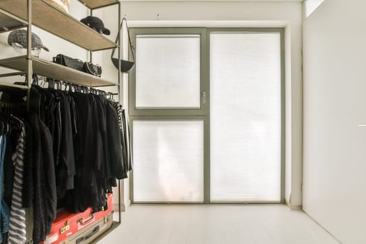 A tidy and organized minimalist closet features shelves for hats and a collection of clothing hung neatly. Natural light filters through the window, enhancing the space.