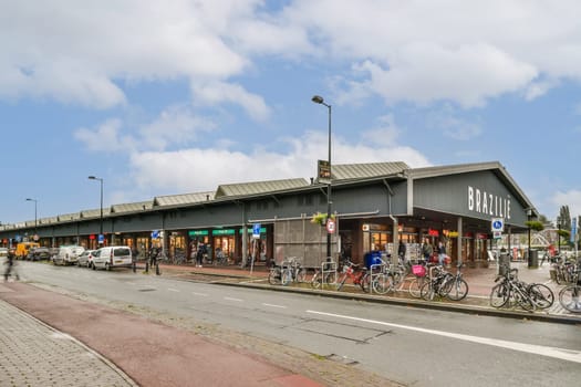 A bustling modern shopping area featuring a variety of stores, outdoor seating, and parked bicycles along a well-paved street under a clear blue sky.