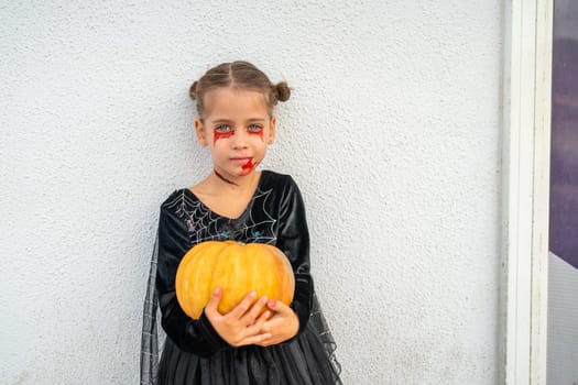 Girl in Halloween makeup dressed in black costume with spiderweb details holding pumpkin while standing against wall