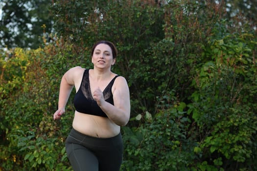 Overweight woman with cellulite in black sports bra running in park against green background, fast running, dynamics.