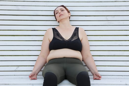 Fat woman with cellulite is tired after fitness. Woman sitting on a white bench in a relaxed pose.