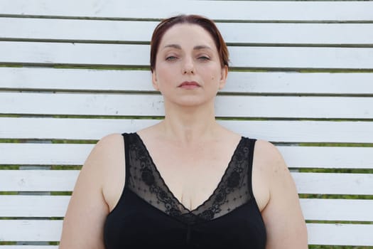A woman in a black bra sits against a white plank wall, looking at the camera.