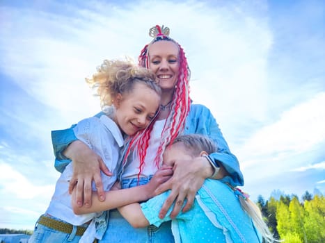 A mother joyfully holds her two daughters close, sharing smiles and laughter on a warm, sunny day outdoors.