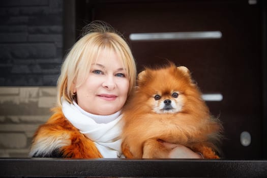 A woman with blonde hair lovingly carries her fluffy red Pomeranian in the winter season against a stylish backdrop.