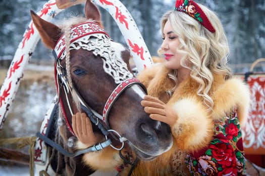 A Russian girl in a vibrant national dress interacts with a horse, celebrating Maslenitsa in a snowy landscape.