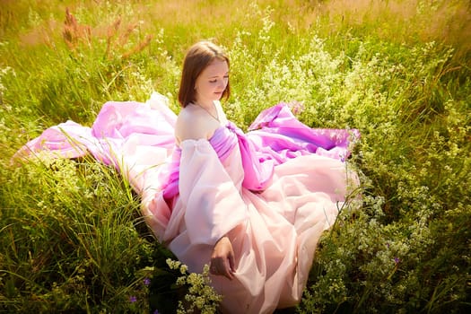 A girl with shoulder-length hair relaxes in a vibrant pink dress amidst lush green grass and blooming wildflowers under a clear blue sky.