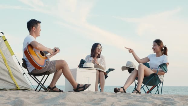 Happy friend have fun playing guitar and clap in camp they smiling together in holiday on sand beach near camping tent vacation time at sunset, Young Asian group woman and man in summer travel outdoor