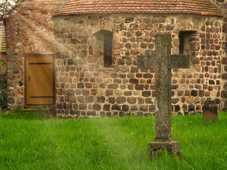 A historic stone cross in front of an old rustic church with small windows. Lively green grass in the foreground adds depth and contrast to the picture.