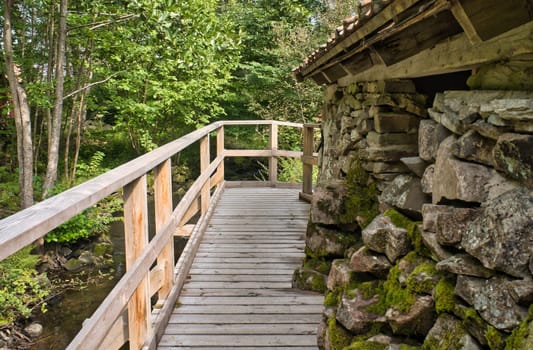 Old watermill surrounded by green nature. A small stream flows over moss-covered stones, surrounded by trees. Idyllic landscape with rustic wooden architecture.