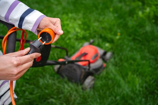 A person is connecting an electric cord to the lawn mower in a lush green yard. The scene captures the vibrant energy of gardening on a bright, sunny day.