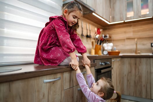 In a cozy and stylish kitchen, two sisters engage in playful antics. One sister sits on the counter while the other pulls on her legs, both laughing joyfully.