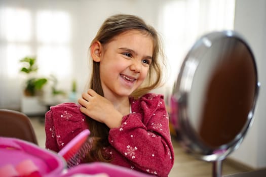 A cheerful young girl smiles while adjusting her hair in front of a small mirror.