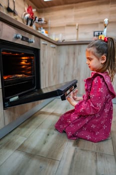 A young girl with long hair, dressed in a vibrant pink dress, kneels in front of a modern oven, eagerly watching as the door opens.