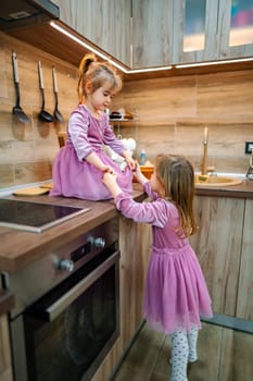 Two young sisters enjoy a playful moment as one stands on a kitchen countertop, adjusting their lovely pink dresses. Laughter fills the air in this inviting, sunlit space adorned with wooden finishes.