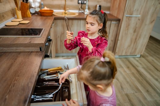 In a beautifully designed kitchen, two girls examine a drawer overflowing with utensils. Their curiosity sparks laughter and excitement as they uncover hidden treasures within the culinary space.