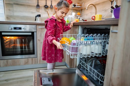 In a bright and airy kitchen, a little girl in a charming red dress joyfully assists with chores by carefully placing vibrant dishes into the open dishwasher amidst a cozy home atmosphere.