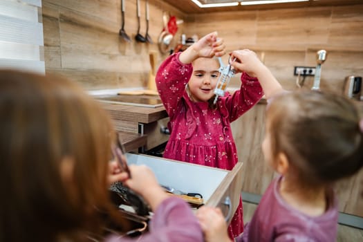 Three children gather around a kitchen drawer, filled with various utensils. One child excitedly holds up a measuring cup, sharing the fun of discovery while the others watch with eager anticipation.