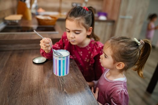 In a warm and inviting kitchen, two young girls share a delightful experience as they savor ice cream from a colorful container on a wooden table, filling their time with laughter and joy.