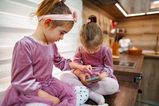 In a warm, inviting kitchen, two girls in matching dresses engage with a smartphone, sharing laughter and excitement. Natural light illuminates their joyful faces as they bond over games.