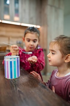 Two young girls engage in a playful activity, carefully mixing a colorful substance from a container. The warm kitchen setting enhances their joyful interaction as they explore creativity together.