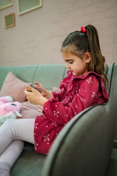 A girl sits comfortably on a modern sofa, engrossed in her play; her adorable outfit and the warm decor create a charming ambiance perfect for relaxation and creativity.