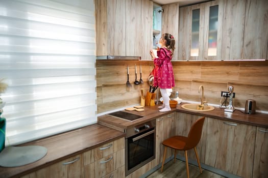 In a cozy kitchen with wooden cabinetry, a small girl in a pink dress stands on the counter, reaching up with curiosity. Sunlight streams in, adding warmth to the playful atmosphere.