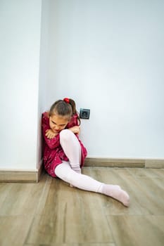 In a quiet room, a young girl huddles in the corner, wearing a playful red dress and pink tights. Her expression reveals a mix of deep thought and a desire for comfort.