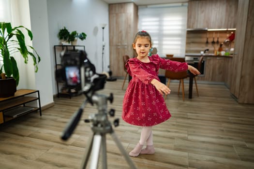 A young girl in a vibrant dress showcases her dance moves in a warm, inviting living room. Bright light fills the area as she captures the joy of creativity on camera.