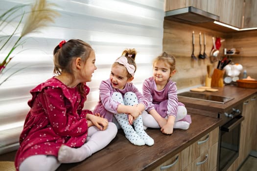 Three young sisters sit on a kitchen countertop, engaging in playful conversation. Dressed in charming outfits, they radiate joy in their cozy, well-lit space filled with warmth and laughter.