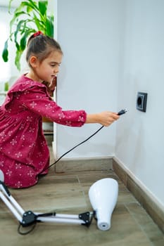 A child dressed in a vibrant red dress is focused on inserting a plug into a wall socket. Sunlight floods the contemporary room, highlighting her concentration.