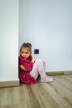 A young girl in a vibrant red dress sits curled up in a corner, her expression deep in contemplation as she gazes at the wall. The serene indoor setting enhances her solitude.