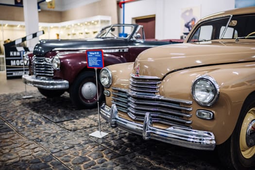 Shiny beige Pobeda car standing in front of a vintage maroon car in a museum or exhibition
