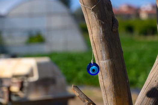 A vivid blue amulet dangles from a weathered wooden post, swaying gently against a lush green field backdrop. The sun casts a warm glow on the serene landscape, creating a peaceful atmosphere.