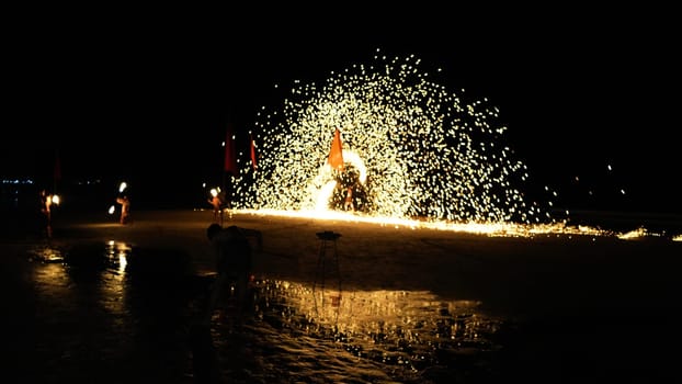 Fire dancers performing a show on a beach at night, creating a shower of sparks, with other fire dancers in the background and a photographer capturing the scene