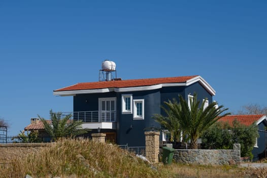 A contemporary home showcases elegant architecture with a vibrant red roof and surrounding palm trees. The serene location evokes tranquility on a sunny day.