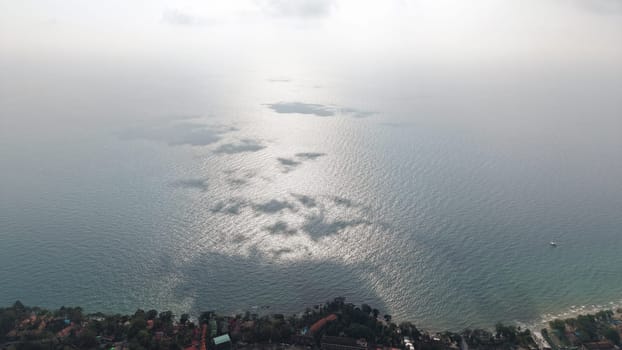 Aerial view of sun reflecting on calm ocean water, creating a mesmerizing pattern of light and shadow, with a coastal village visible at the bottom and a layer of haze obscuring the horizon
