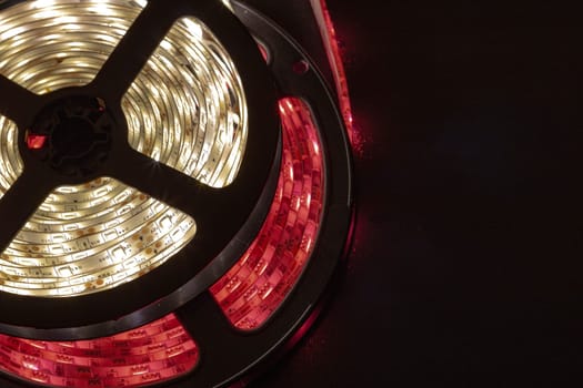 Detailed photo of LED strip lights rolled on a black reel, combining warm white and red illumination. Ideal for illustrating technology, modern lighting, decoration, and electronic components.