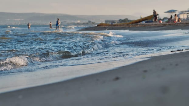 Small waves gently washing sandy beach of Crimea at sunset, with tourists enjoying summer vacation in the background