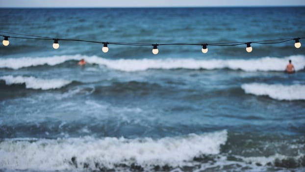 String of lights hanging over the crimean sea at dusk while tourists swim and enjoy the waves