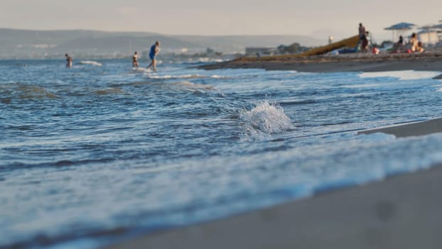 Soft waves gently lapping the sandy shore of a crimean beach as the sun sets, creating a tranquil scene with blurred tourists enjoying the summer evening