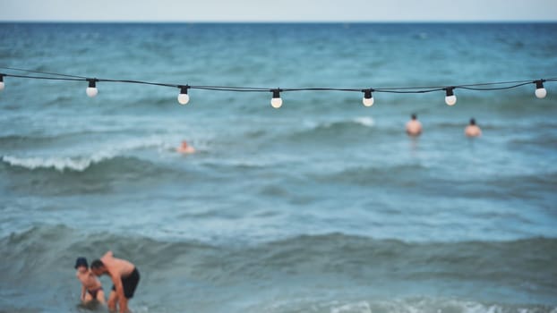 Father and son bathing in the sea in Crimea, Russia, with string lights and tourists in the background