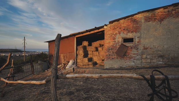Bales of hay stacked inside a stable in Crimea, near Belogorsk, with horse harness hanging on a wooden fence at sunset