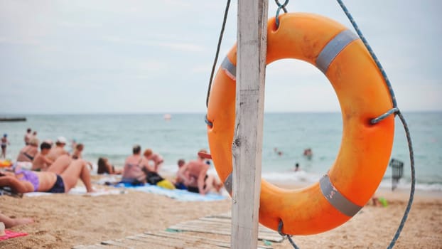 Orange lifebuoy hanging on a wooden pole on a beach with tourists relaxing on the sand in Koktebel, Crimea